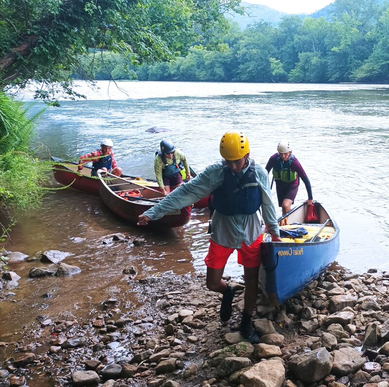 A group of people are preparing to go canoeing on a river. They are wearing helmets and life jackets for safety. The canoes are colorful, and the river is surrounded by lush greenery. The people seem to be enjoying the outdoors and are ready for a fun adventure on the water. The rocky shore suggests a natural and scenic environment.